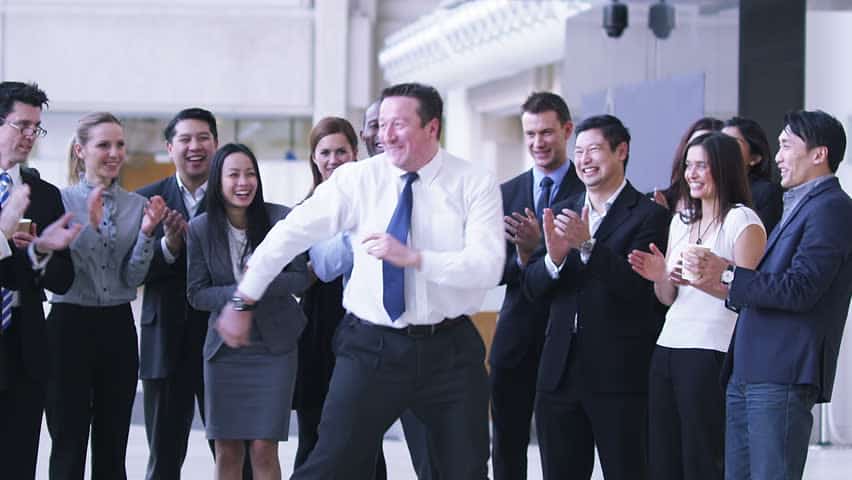 Man in a blue tie dancing and surrounded by his clapping and happy officemates